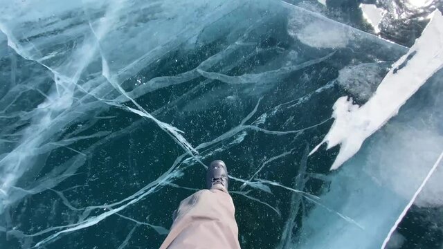 A man walks on frozen Lake Baikal with blue transparent ice with deep cracks. Beautiful winter landscape with clean, smooth ice. Natural landmark of Russia. Top view of the legs.