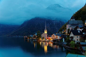 Fototapeta na wymiar Morning view of Hallstatt, a peaceful lakeside village in Salzkammergut region of Austria, with majestic mountains & lights of village houses & the church reflected on lake water in a deep blue mood