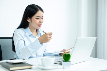 Asian woman working on a laptop with a cheerful and happy smile while working and take notes at the office