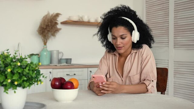 Cute African American woman in big white headphones listens to music with pleasure using smartphone. Black woman posing while sitting at table the bright kitchen. Slow motion ready, 4K at 59.97fps.