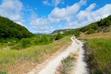 Fototapeta na wymiar Mountain road with green meadows and mountain peaks under the blue sky with clouds. The Crimea, Mangup Kale.