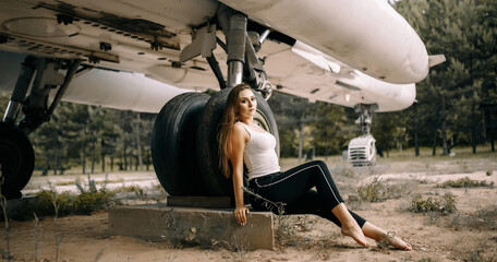 beautiful young brunette girl sits on the background of old military aircraft. Girl in a white shirt and black pants in nature. Military equipment. Portrait half-length. girl posing