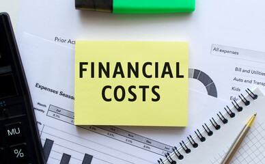 Text FINANCIAL COSTS on the page of a notepad lying on financial charts on the office desk.