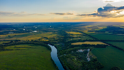 Fototapeta premium Aerial view of a beautiful summer hilly landscape with a river while sunset. Wide green valley with a river running in the middle. Green meadows. Top view over beauty nature.