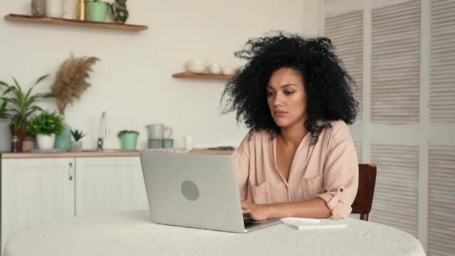 Cute African American woman studying online or remote working using laptop while taking notes in notebook. Black female posing sitting at a table in bright kitchen. Slow motion ready, 4K at 59.97fps.