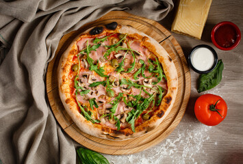 Homemade pizza with arugula, pickles, ham and sauce on a wooden pizza plate.