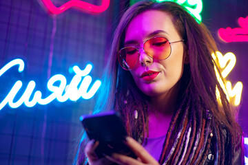 Fashion girl using her smartphone while standing near neon signs in nightclub