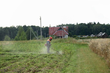 Fototapeta premium A worker man in hard hat helmet in coveralls mow the grass with a motor trimmer on a green lawn on rural house and forest background at summer day, countryside work view