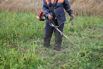Caucasian farmer worker in overalls mows the grass with a motoring trimmer in the field close-up on a summer day - season working, landscaping