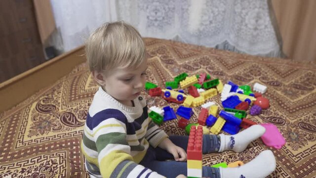 The child is played with toys. Little boy assembles the constructor on the bed. Multi-colored toys. Small baby details. Self-isolation of the baby in the house. Colored children's socks and tights