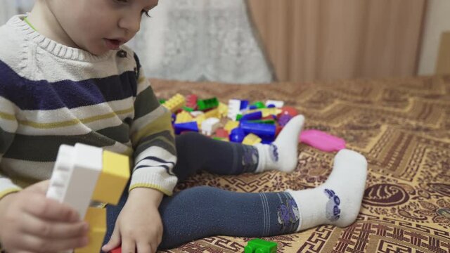 The child is played with toys. Little boy assembles the constructor on the bed. Multi-colored toys. Small baby details. Self-isolation of the baby in the house. Colored children's socks and tights