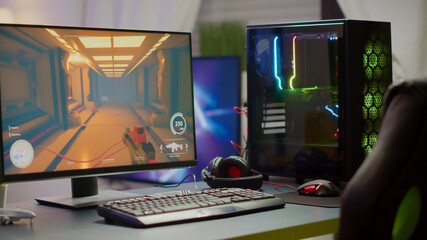 RGB powerful personal computer with first-person shooter game on screen in gaming studio with no people at desk. Empty cozy room with modern design is lit with warm and neon light.