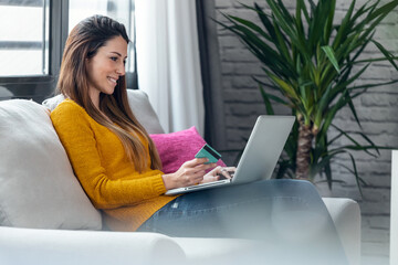 Smiling young woman paying something online with her credit card in laptop while sitting on couch at home.