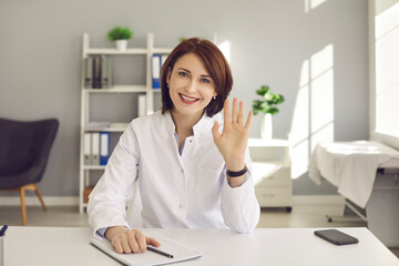 Webcam view. Friendly middle-aged female doctor waving hand greeting a patient via video link. Woman in a medical gown sits at her workplace and provides online consultations to patients.