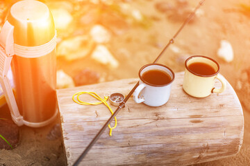 a thermos and mugs are on the shore, next to a compass and a fishing rod. The concept of fishing and outdoor recreation.