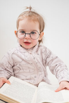 Cute toddler girl in glasses reading book on white background
