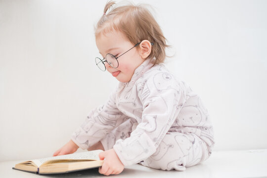 Cute toddler girl in glasses reading book on white background