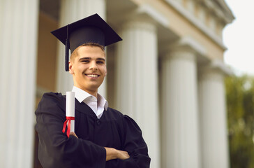Happy male college graduate student in mortatboard holding diploma near university campus outdoors,...
