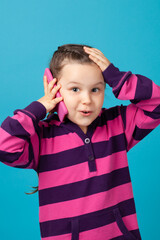 vertical close-up portrait of a surprised, shocked, delighted five-year-old girl making a phone call and holding her head with her hand isolated on a blue background.