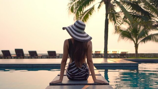 Sexy woman sitting on the edge of the swimming pool at an exotic hotel in Florida wearing botched spotty monokini and striped hat looking at sunset beach, back view slow-motion handheld
