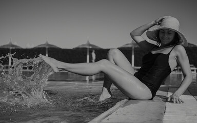 An attractive girl sits near the pool in a swimsuit and a straw hat.