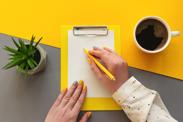 Female hands with writing on paper. Office table desk trendy yellow and grey colors. Workplace with...