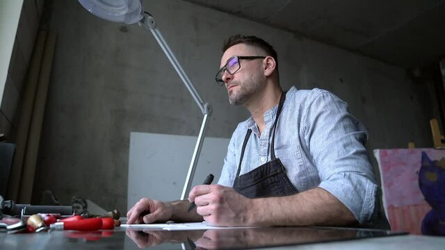 Lithograph artist making handmade stamp. Linocut artist printmaking manufacture work. Bearded middle aged man in apron making sketch on linoleum. 4k footage