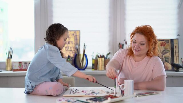 Mother and daughter painting together at home with paintbrushes and watercolors. Young woman sitting and helping or teaching daughter to draw. 4k footage