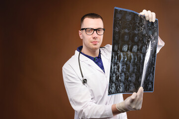 A young male doctor in glasses with a stethoscope around his neck holds an X-ray image of the spine...