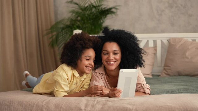 African American woman and litle girl talking on video call using digital tablet. Mother and daughter posing, lying on the bed in a bedroom with a bright interior. Slow motion ready, 4K at 59.97 fps.