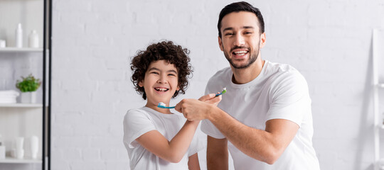 Positive arabian father and son holding toothbrushes and toothpaste, banner
