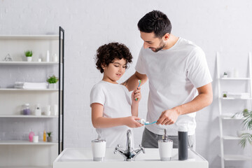Fototapeta premium Smiling muslim boy holding toothbrush and toothpaste near father in bathroom