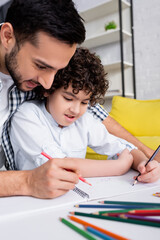 Naklejka premium happy arabian dad and son drawing with pencils in sketchbook together on blurred foreground