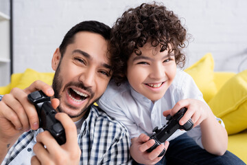 KYIV, UKRAINE - JANUARY 15, 2021: astonished muslim man playing video game with cheerful son at home