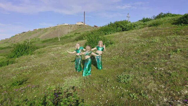 Aerial view from a drone flying over three European women in exotic oriental glittering costumes dancing oriental dances and blowing kisses against the backdrop of green tundra. The camera moves away