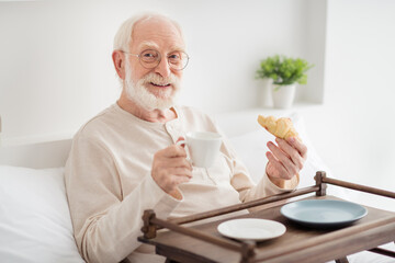 Photo of elderly man pensioner happy positive enjoy morning breakfast on bad home drink tea eat croissant