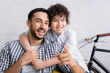cheerful arabian boy embracing laughing father pointing with finger at camera