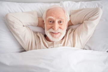 Photo of cheerful positive mature man nightwear arms behind head lying bed having rest indoors...