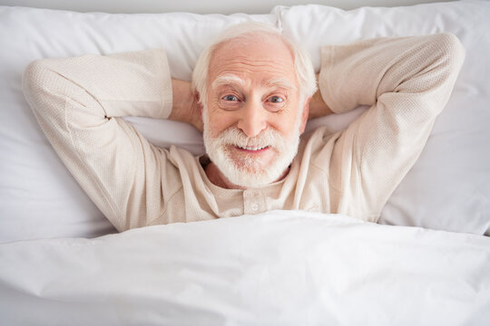 Photo of cheerful positive mature man nightwear arms behind head lying bed having rest indoors inside room home