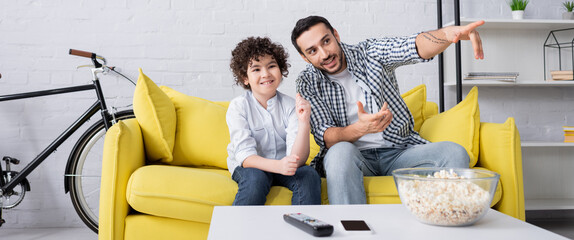 cheerful arabian man pointing with fingers while watching movie with son, banner