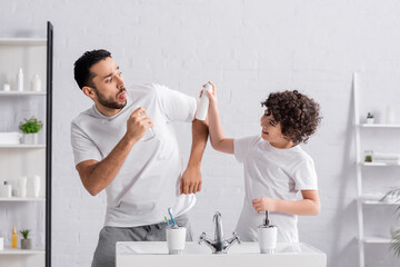 Smiling Arabian son holding deodorant near father with sprayer and sink