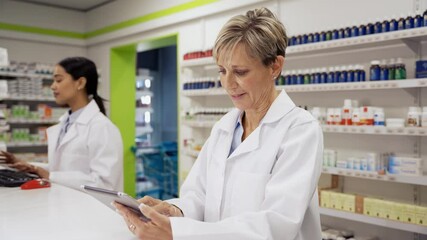 Caucasian female pharmacist wearing coat scrolling to find scripts on digital tablet in pharmacy 
