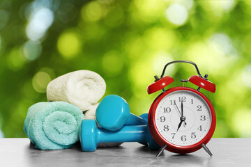 Alarm clock, towels and dumbbells on marble table outdoors. Morning exercise