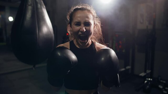 Portrait of female boxer standing at dark gym, looking intensely at the camera and screaming