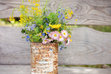 a bouquet of wildflowers of forget-me-nots, daisies and yellow dandelions in full bloom in a rusty rustic jar against a background of wooden planks in nature. cottagecore scene