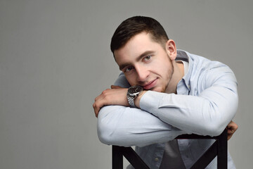 Pensive brunette man in blue shirt sitting on the chair and looking at camers over grey background