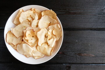 Chips of fresh apples with cinnamon are on a white plate on a background of black wooden boards. Organic apple chips. Dried fruits. Healthy sweet snack. Dehydrated and raw food. Copy space.