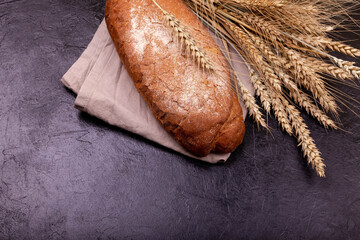 Cropped view of brown loaf on stone black surface. Rye bread on dark background. Rustic sourdough bread with crispy crust. Loaf of whole grain bread and shocks of wheat. Freshly baked homemade bread.