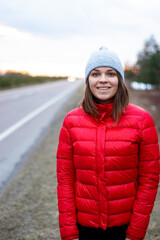 smiling young brunette woman standing on the road at sunset in a red jacket