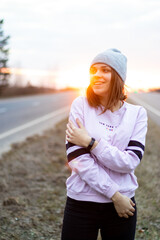 woman standing on the road at sunset. Woman in a hat, pink jacket, the sun would be in the back.